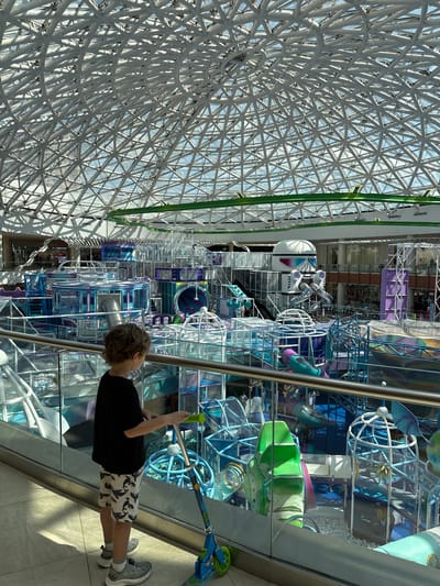 A young boy stands on a tiled floor, looking towards an elaborate indoor playground with a large, geodesic dome ceiling. He is wearing a black t-shirt, patterned shorts, and sneakers, and is holding onto the handlebars of a blue scooter. The playground features multiple levels of tubes, slides, climbing structures, and ball pits, with a predominantly blue and white color scheme. In the background, shops and other parts of the indoor space are visible. The lighting suggests it is daytime, with sunlight filtering through the transparent dome. The provided image does not contain any information that would suggest this location is in the city of Sovkhoza imeni Lenina, Russia; the scene appears to be a modern indoor entertainment facility.
