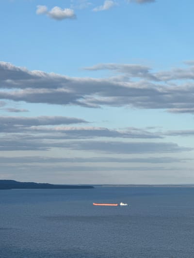 A vertical shot depicts a body of water, presumably a lake or ocean, occupying the right half of the frame. A solitary boat is visible on the water's surface, trailing an orange object behind it. Adjacent to the water, on the left, is a landmass, potentially a shoreline or an island. The sky above is filled with diverse cloud formations, ranging from wispy, linear clouds to larger, more textured cloud masses. The overall color palette is dominated by blues and grays, with a hint of orange from the object trailing the boat. The location is presumed to be near Galevo, Russia, based on the prompt.