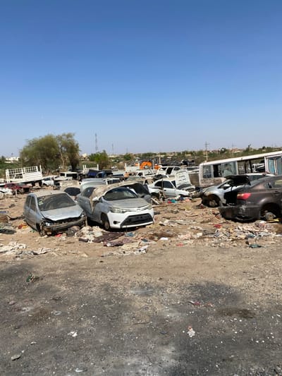 The image shows a large junkyard in Tayba Alhasanab, Sudan (latitude: 15.36, longitude: 32.528), littered with discarded vehicles and debris. There are at least a dozen cars, many of them damaged or missing parts, along with a bus, a truck, and various scattered pieces of metal, fabric, and plastic. The vehicles are arranged haphazardly, and there is no sign of any people present in the scene. The ground is covered in dust and dirt, and the sky is a clear blue.