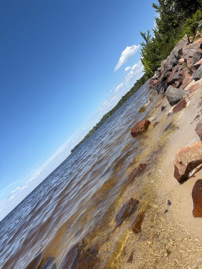 The image depicts a natural scene at the Kyiv Reservoir near the village of Kozarovychi, Ukraine. The foreground is dominated by a shoreline where calm, brown water gently laps against a sandy beach dotted with reddish-brown rocks. The water is shallow enough to reveal the sandy bottom in places. Beyond the immediate shoreline, the water stretches to the horizon, meeting a distant treeline on the left side of the frame. The sky is a vibrant blue, with scattered white clouds adding texture. The perspective is tilted, giving the impression of looking out over the water from a low angle.