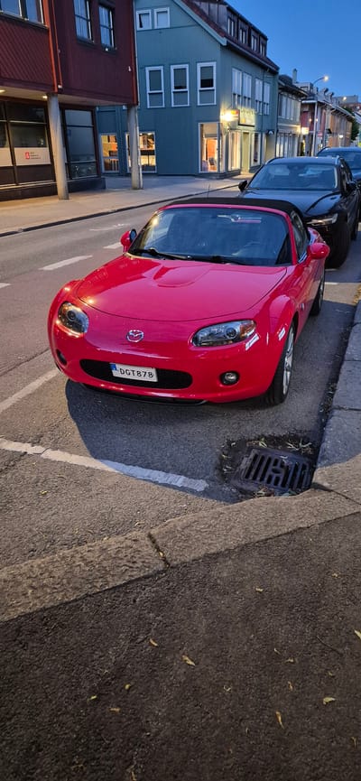 A red Mazda MX-5 convertible is parked on a street in Lillehammer, Norway. The car's license plate reads "DGT 878". It is parked next to a black SUV and in front of a building with a blue facade and several windows. The street appears to be residential or commercial, with sidewalks and a storm drain visible. The lighting suggests it is either early morning or late evening. No people are visible in the image.