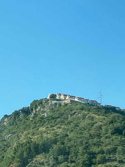 The image shows a hillside in Blizikuće, Montenegro, on a clear day. The hillside is covered in green vegetation and has buildings constructed along its slope. Power lines run along the side of the hill, and an electrical tower is visible in the distance. The sky is clear blue.