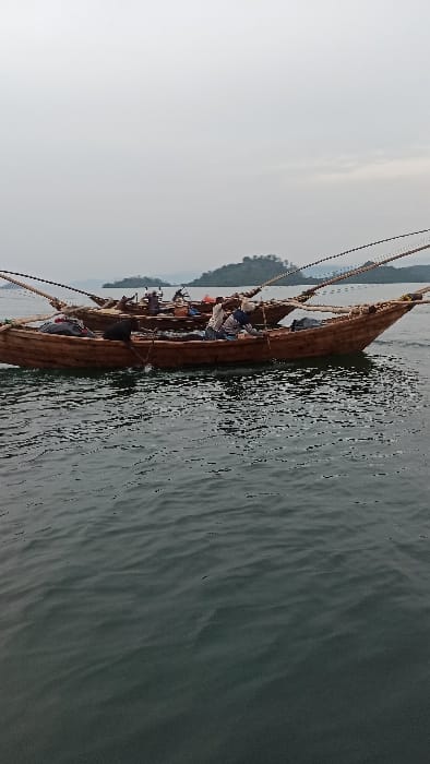 The image, rotated 90 degrees clockwise from its intended orientation, captures a long, narrow wooden boat on a body of water, likely a lake or wide river, off the coast of Bwishyura, Rwanda. Several people are seated within the boat, suggesting travel or a group activity. The individuals appear to be wearing casual clothing. The boat itself is rustic, made of dark wood, and has ropes running along its length, possibly for steering or securing items.
The water around the boat is dark and shows gentle ripples, indicating either slow movement or a calm surface. In the background, a distant landmass, possibly hills or mountains covered in vegetation, rises above the water. The sky is bright but somewhat hazy or overcast, suggesting daytime, possibly morning or late afternoon, with soft, diffused light. No specific activity beyond being on the boat is clearly discernible, nor are any strong emotions expressed by the individuals visible. There is no visible text in the image.