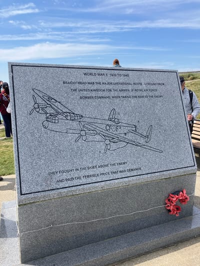 The image shows a memorial stone dedicated to the airmen of the Royal Air Force Bomber Command who fought during World War II from 1939 to 1945, located in Eastbourne, United Kingdom. The stone is gray with black lettering and features an engraved outline of a bomber plane with the letters CFOF2 on it. The inscription reads: "WORLD WAR II 1939 TO 1945. BEACHY HEAD WAS THE MAJOR OPERATIONAL ROUTE OUTBOUND FROM THE UNITED KINGDOM FOR THE AIRMEN OF ROYAL AIR FORCE BOMBER COMMAND, WHEN TAKING THE WAR TO THE ENEMY. THEY FOUGHT IN THE SKIES ABOVE THE ENEMY AND PAID THE TERRIBLE PRICE THAT WAR DEMANDS." On top of the memorial stone, there is a wreath of artificial poppies. People are visible in the background, including one sitting on a bench. The sky is blue with streaks of white clouds.