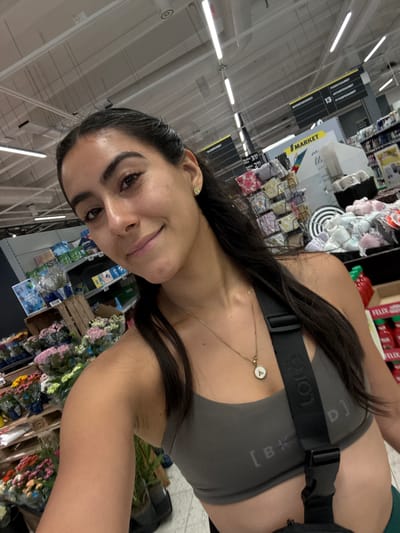 The image captures a selfie of a young adult female inside an S Market supermarket in Imatra, Finland. The woman, with dark hair, dark eyes, and a medium-dark complexion, smiles subtly at the camera. She wears a dark olive-green sports bra or crop top, a golden necklace with a circular pendant featuring a white center and a small dark letter 'A', and a small white daisy earring in her right ear. A black strap, likely from a bag, with "LOI" and "[BK_LD]" embossed text, diagonally crosses her chest. The supermarket environment is brightly illuminated by numerous overhead fluorescent strip lights on a white ceiling with visible ventilation ducts. Hanging signs, dark grey with white Finnish text, indicate product sections such as "TALOUSPAPERIT" (household paper), "PUHDISTUSAAINEET" (cleaning agents), "ASTIANPESUAINEET" (dishwashing detergents), and "PYYKINPESUAINEET" (laundry detergents), with an aisle number "13" also visible. A prominent white sign displaying a yellow "S MARKET" logo is in the background. To the left, a diverse selection of flowers in various colors (pink, blue, yellow, red) is arranged on wooden pallets. To the right, displays include packaged goods, a basket of folded white and light purple garments, and a stand featuring red bottles of "FELIX" brand products. The floor is covered with light-colored tiles.