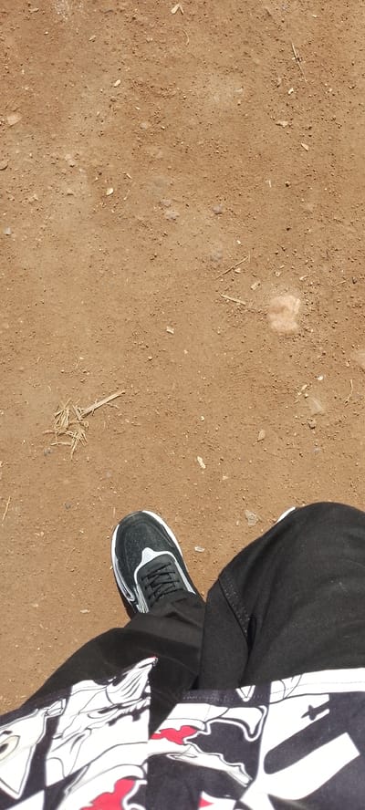 The image shows a close-up, overhead view of a person's legs and feet on a dusty, dirt ground. The person is wearing black pants and a black and white sneaker with a white sole and white accents on the side. The ground is a light brown color and appears dry and uneven, with small pebbles and some scattered dry grass. The lower portion of the image reveals a patterned fabric, likely from the person's clothing, which is black with a white design featuring red accents. There are no discernible city landmarks or indications of Ajio, Nigeria.