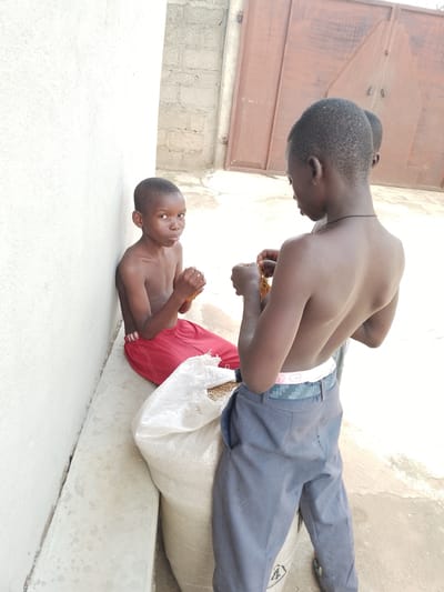 Three young boys are shown outdoors in what appears to be a casual, urban environment in Balewa, Nigeria. A boy in red shorts is seated against a white wall, holding something in his hands. To his right, another boy in gray pants stands and examines something in his hands, which appears to be food items. A third boy's head is visible behind the standing boy, partially obscured. A large, off-white sack is positioned in front of the seated boy, containing grain. The background features a rough, textured wall and a large, reddish-brown metal gate. The ground is paved with concrete.