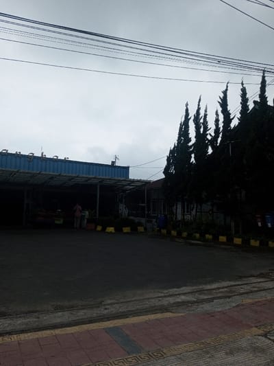 A cloudy day shows a roadside scene. A long, blue building with a sign that reads "Angkhar" is visible with an open storefront. A person stands near the building. Tall, dark green trees line the right side of the building. The foreground is a street and sidewalk with patterned tiles. Overhead are visible utility lines.
