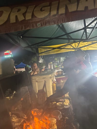 A food stall with open flames and smoke displays a grill piled with bread rolls. A person tends the grill. A woman is visible inside the stall's covered area, seemingly part of the staff. Signage and lettering are evident on the stall's exterior, suggesting a commercial establishment. A tent structure frames the stall, and a brightly lit sign is visible in the background.