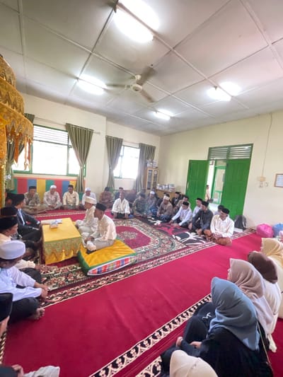This image captures an indoor gathering, likely a religious or ceremonial event, in a room in Tibang, Indonesia. The room features a red carpet covering most of the floor, with decorative patterned borders. In the foreground, several people are seated on the carpet, many wearing traditional Islamic attire, including white peci (caps) and long white shirts. To the left, a decorative golden structure, possibly part of an altar or stage, is visible.

A group of men are seated in a semi-circle around a low, yellow-covered table in the center of the room. Some men are engaged in conversation, while others appear to be listening or observing. The room has large windows with green curtains and is illuminated by overhead lights. In the background, more people are seated on the floor. There are also green double doors open to another area. The overall atmosphere suggests a formal or communal gathering.