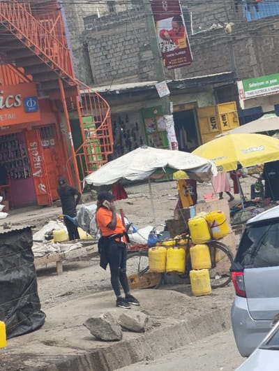 The image depicts a bustling commercial street scene in Matungulu west ward, Kenya. In the midground, a woman in an orange t-shirt, black cap, and black pants is standing on an unpaved surface, holding a dark-colored mobile phone to her right ear. To her immediate right, a bicycle is heavily loaded with at least eight large yellow jerrycans, suggesting a water or liquid vending operation. Two irregular grey rocks are situated on the ground in front of the woman. Behind her and to the right, a white market umbrella is open, alongside a bright yellow umbrella with "Belikal" and "WIN" branding. Underneath and around these umbrellas, indistinct figures and market goods are visible, including one person in a pink garment. To the left, a man in a black shirt stands near a wooden market stall with white items. A black tarp covers an object on the far left. On the right, the rear section of a light-colored vehicle is partially visible.
The background features several multi-story buildings constructed of rough brick and concrete. Prominent signage includes an "Optics" store, "Angels" (possibly a beauty salon or clothing store), a "Chess Classes" banner with contact numbers, and "TIMES ELECTRICAL & HARDWARE" with a "Safaricom m-pesa" logo. An external orange metal staircase is affixed to the left building, and a green spiral staircase is visible further back. Overhead utility lines span the scene. The ground is largely unpaved dirt and gravel, with a defined curb separating it from what appears to be a street, parts of which are also unpaved.