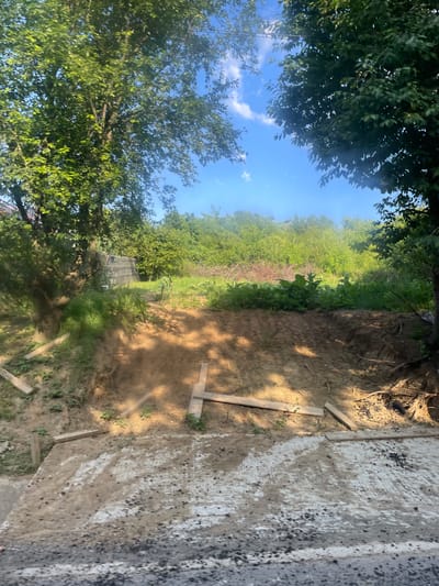 The image shows an outdoor scene with lush green trees and vegetation under a bright blue sky with scattered clouds. In the foreground, there is a patch of disturbed earth, possibly indicating recent construction or landscaping. Several wooden planks are visible, some lying on the ground and others partially embedded in the soil, suggesting some form of construction or a makeshift path. The area appears to be a natural, undeveloped landscape, perhaps a yard or a rural area. No people or specific man-made structures are clearly identifiable beyond the wooden planks and what appears to be a large black tarp in the background, possibly covering something or part of a structure. The overall impression is of a sunny, natural environment in the process of being worked on or cleared.