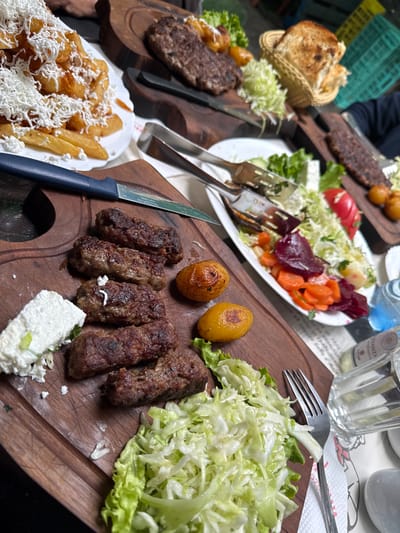 A table is set with an assortment of food in an outdoor setting. A wooden cutting board holds ćevapi (grilled minced meat) accompanied by cooked potatoes, crumbled white cheese, and shredded lettuce. Other dishes include bread in a basket, a burger with a potato, a salad with tomatoes, carrots, and beets, and a plate of french fries topped with crumbled cheese. A bottle of water and glasses are also visible on the table. The table is covered with a tablecloth, and cutlery is placed around the dishes. The location appears to be Strumica, North Macedonia, based on the food typically found there. At least one person is seated at the table, visible by their arm.