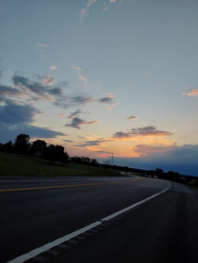 A vertical, low-angle shot captures a desolate highway at dusk, with a gradient sky transitioning from pale blue to the warm hues of sunset. The sky is adorned with scattered clouds, some tinged with pink and orange, suggesting the last moments of daylight. The road, paved and dark, curves gently to the right, delineated by double yellow lines in the center and a dashed white line on the shoulder. A small, indistinct structure, possibly a house, is nestled amongst dark silhouettes of trees on a grassy incline to the left of the road. Two vehicles are visible in the distance, driving away from the camera on the curving road, their headlights faintly illuminated. The overall impression is one of quiet solitude and the beauty of twilight.