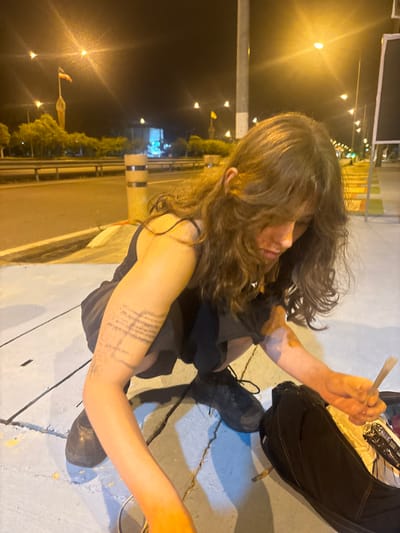 The image depicts a night scene on a concrete sidewalk next to an asphalt roadway in Thap Sakae, Thailand. A young individual with long, wavy brown hair, wearing a dark tank top and dark shorts or skirt, is squatting or crouching. Their attention is directed downwards toward objects on the ground.

On the individual's left upper arm, a faded tattoo or drawing of geometric lines is visible. Their right hand holds a hand-rolled item resembling a cigarette or joint. An open, dark-colored bag, possibly a backpack, rests on the ground beside the individual, with some light-colored paper and other indeterminate items visible inside.

The immediate environment includes a cracked concrete sidewalk with yellow markings and the adjacent paved roadway. Cylindrical black and light-colored traffic barriers are positioned along the edge of the road. In the background, multiple bright streetlights illuminate the area. Further in the distance, two tall flagpoles with flags, exhibiting colors consistent with the Thai national flag, are discernible. Other blurred lights and structures indicate an urban setting at night.
