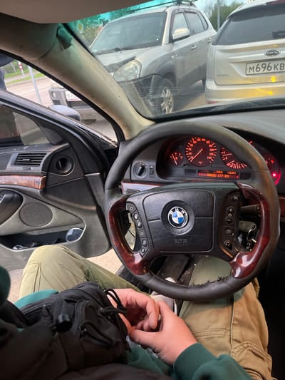 The image depicts the interior of a car, focusing on the dashboard, steering wheel, and a portion of the driver's area. The steering wheel is a BMW model, as indicated by the emblem. The dashboard includes a speedometer, fuel gauge, and other instruments. A Ford is visible through the windshield. The license plate of the Ford reads "M696 KB". The image was taken in the city of Вёшки, Russia. The driver is wearing a green sweatshirt.