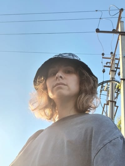 In Maslova Pristan', Russia, a low-angle, medium shot captures a person wearing a dark bucket hat and a grey t-shirt against a bright blue sky. The person's face is partially illuminated by sunlight, highlighting their light brown, shoulder-length hair. Behind them, a utility pole with electrical wires and insulators is visible, suggesting an outdoor, possibly urban or semi-urban, setting. The lighting indicates it is daytime, with the sun positioned behind the person and to their left, creating a lens flare effect.