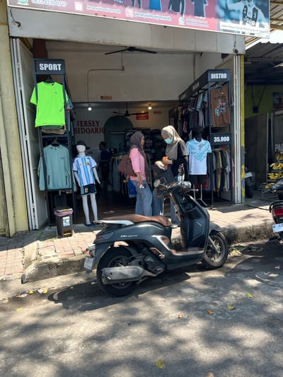 The image depicts a street-level view of a storefront in Sepande, Indonesia, selling sportswear and apparel. The storefront is open to the street, with clothing racks visible both inside and outside. Several people are present in front of the store, appearing to be customers and staff. One woman, wearing a headscarf and face mask, is interacting with another person, possibly a customer. Another woman in a pink shirt and jeans stands to the left, holding a bag. A mannequin dressed in a soccer jersey is displayed outside. A black scooter is parked in the foreground, casting a shadow on the paved ground. The environment appears to be a typical commercial street with buildings lining the road. The overall impression is that of a lively local business operating in a warm, sunny climate.