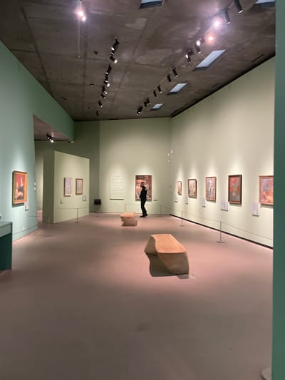 The image depicts the interior of an art gallery, identified as the Ordrupgaard Museum in Klampenborg, Denmark. The spacious room features pale green walls and an exposed grey concrete ceiling fitted with black track lighting fixtures and several recessed square skylights. The floor is covered in a uniform light taupe material. Numerous framed paintings of various sizes and styles are displayed on the walls, individually illuminated by spotlights. A single individual, dressed in dark clothing, stands in the mid-ground, facing and observing a painting on the far wall. Two light-colored, curved benches are positioned on the floor in the foreground and mid-ground. Stanchions with ropes delineate a viewing area along the right wall. An adjacent gallery section featuring additional artworks and a display case is visible on the left side of the frame.