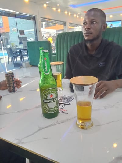 A man is seated at a table in a restaurant in Ujevwu, Nigeria, at the coordinates 5.472 latitude and 5.818 longitude. He is wearing a dark blue polo shirt and has short black hair. The man is looking to the right of the image. He is holding a glass of beer in his right hand, resting his left hand on the table. The table is white marble with a gold trim. In front of him is a bottle of Heineken beer, a glass of beer with a coaster, and a card with a QR code that says "Scanase" and "to only Deluxe account." The restaurant has a green, leather booth behind the man and large windows with a view of an outdoor area.