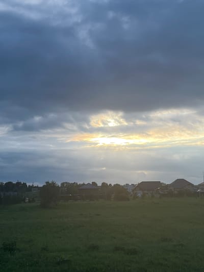 This is a vertical, wide-angle photograph of a suburban landscape at sunset. The sky dominates the left half of the frame, filled with dramatic clouds ranging from dark, ominous grey on the far left to lighter, wispy formations illuminated by the setting sun on the right. The sun itself is a bright, almost blinding glow peaking through the clouds, casting a warm, golden light that reflects off the cloud edges.
To the right of the sky, a line of houses with peaked roofs is visible, nestled amongst trees. The houses appear to be typical suburban dwellings, and their architecture suggests a European or North American setting, consistent with the Kamskiye Polyani location. The trees are leafy and green, indicating a summer or late spring season.
The right half of the image is a large, open field of green grass. The grass is somewhat uneven and appears to be a park or undeveloped lot. The overall impression is one of a peaceful, end-of-day scene in a quiet residential area. The lighting suggests it is late evening, around sunset, with a mix of cloudy and clear skies. There is no visible text in the image.