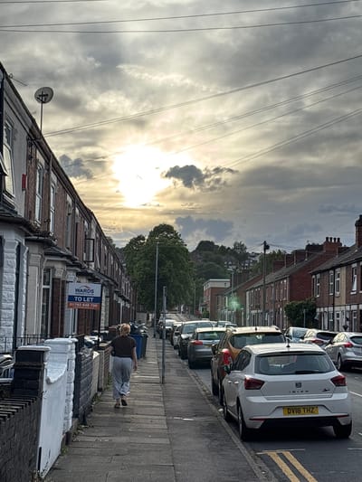 A street scene in Stoke-on-Trent, United Kingdom, shows a row of terraced houses lining a paved sidewalk. Parked cars are visible along the right side of the street, with license plates visible. An adult female walks along the sidewalk, facing towards the distant end of the street. A 'To Let' sign from a property management company (WARDS) is affixed to one of the houses. Overhead, power lines stretch across the cloudy sky, where the sun is obscured by cloud cover.