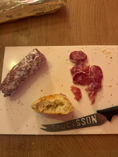 A white cutting board displays a variety of food items. Sliced and whole dry sausage are visible, along with a piece of bread. A knife with the word "SAUCISSON" cut out of the blade is positioned on the board. A sausage wrapper is partially visible to the left of the board. Small black flecks are scattered across the surface. The board is placed on a wooden surface.