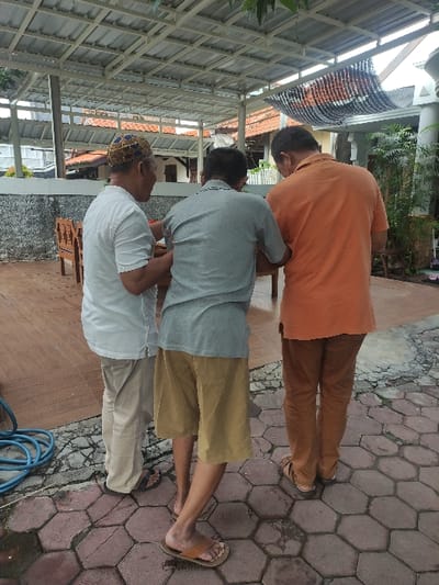The image shows three men standing outdoors on a patterned hexagonal paved surface, likely in Lamongan, Indonesia. The perspective is from behind and slightly to the side of the men.
The man on the right is wearing an orange short-sleeved shirt, brown trousers, and light-colored sandals. The man in the middle has on a grey short-sleeved shirt, mustard or khaki-colored shorts, and sandals. The man on the left is dressed in a white short-sleeved shirt, light khaki trousers, and a patterned head covering, possibly a kufi or peci, which is blue, gold, and white. Their postures suggest they are standing, possibly conversing or looking at something in front of them, though their faces are not visible.
The scene appears to be an outdoor sheltered area, possibly a patio or courtyard, with a metal-framed overhead structure or pergola visible in the upper portion of the image, indicating shade. In the background, there are hints of buildings with reddish-brown tiled roofs and some greenery. A blue coiled hose lies on the paved ground in the foreground on the left side. The lighting suggests it is daytime, with diffused light indicating either an overcast sky or the presence of the overhead structure providing cover. No visible text is present in the image.