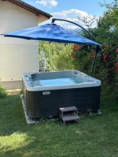 An outdoor jacuzzi is situated in a grassy backyard, partially shaded by a large blue cantilever umbrella. The jacuzzi is dark gray with a white rim and filled with clear water. A small, two-step wooden platform is placed next to it. In the background, a white building with a brown roof is visible, as well as lush green trees and bushes, some of which are flowering with red blossoms. The sky is blue with scattered white clouds, and the overall scene suggests a sunny day. The location is not identifiable as Chavei, Bulgaria based on the provided image.
