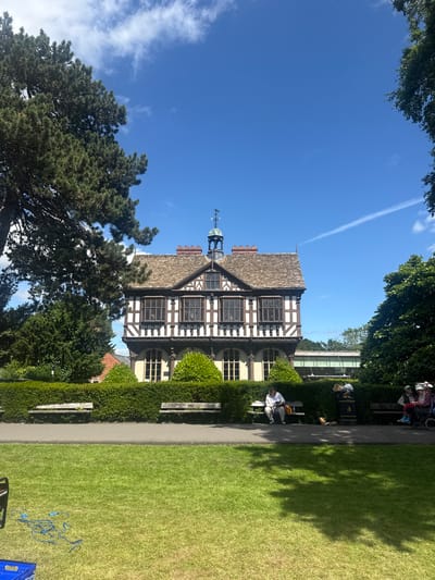 A three-story Tudor-style building with a gabled roof and timber framing stands in the background of a sunlit park. The building's lower level features arched windows, while the upper levels have rectangular windows framed by dark timber. A weather vane adorns the roof's peak. In front of the building, a neatly trimmed hedge separates it from a paved walkway. Several benches are placed along the walkway. On one bench, a person sits alone, looking down at their hands. Further down the walkway, another bench is occupied by two people, one of whom is in a wheelchair. A trash bin stands beside the second bench. The park's foreground is a lush green lawn. To the left, a large evergreen tree partially obscures the building. The sky is a clear blue with a few wispy clouds. The scene is located in Leominster, United Kingdom, at the Leominster Priory.
