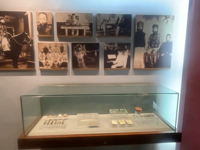 The image depicts a display in a museum, likely located in Hailing District, China, based on the displayed content. A glass display case sits on a dark wooden base. Inside, various artifacts are exhibited. These include what appear to be writing implements (pens or brushes), spectacles, coins or stamps, and possibly seals, each displayed on separate clear plastic platforms. Above the display case, a series of monochrome historical photographs are mounted on the wall. The photographs appear to depict scenes and individuals from Chinese history, featuring portraits of individuals in traditional clothing, group photos, and images of people in various settings. The background wall is a pale blue. A vertical pink wall is visible on the right.