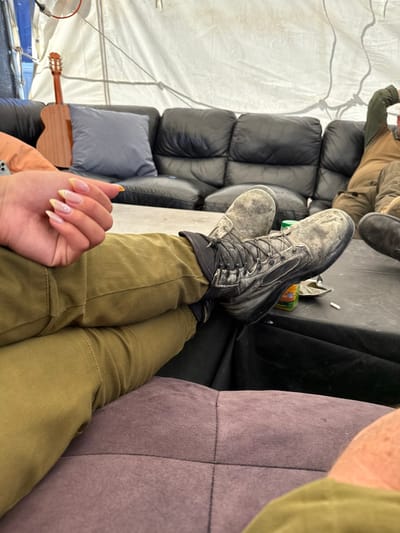The image shows a person wearing olive-green pants and black boots, sitting with their legs resting on a table. The person is wearing nail polish with a yellow hue. Another person, partially visible in the background, is also wearing olive-green pants and sitting on a black leather couch. Both appear to be wearing military-style clothing. A guitar is leaning against a wall, and a can and cigarette are on the table. The scene appears to be inside a tent, possibly a temporary military encampment. The coordinates provided place the scene in the city of Mazkeret Batya, Israel, in a location that may be a military base or training facility.