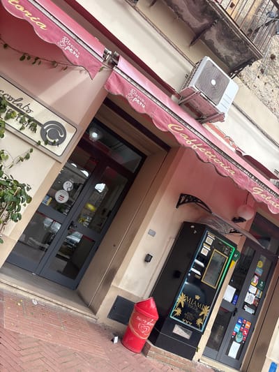 The image captures the exterior of a business in Laureana di Borrello, Italy. The view is taken from below, looking up at the building's facade. A pink awning with the words "Caffetteria Gelato" provides cover above the business entrance. Below the awning, a sign for "Cioccolato" is partially obscured by green foliage. The entrance to the business features glass doors with visible signs for Wi-Fi and Nutella. To the right of the entrance, a "Miami Touch" vending machine is placed. Next to the vending machine is a large, red "Share Happy" ice cream container. An air conditioning unit is mounted on the wall above the awning. The building has a light-colored exterior, and the surrounding environment includes red brick pavement.