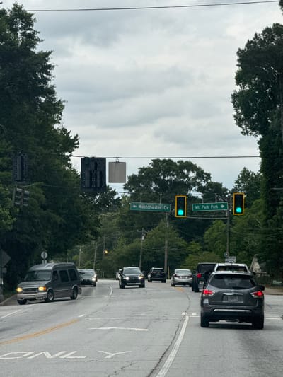 The image shows a street in Lilburn, Georgia, United States. A line of cars is stopped at a traffic light. The street signs read "Westchester Dr" to the left and "Mt. Park Park" to the right. The traffic lights are green. The sky is overcast. Trees are visible on both sides of the street. A white line separates the lane from the shoulder. A speed limit sign is visible.