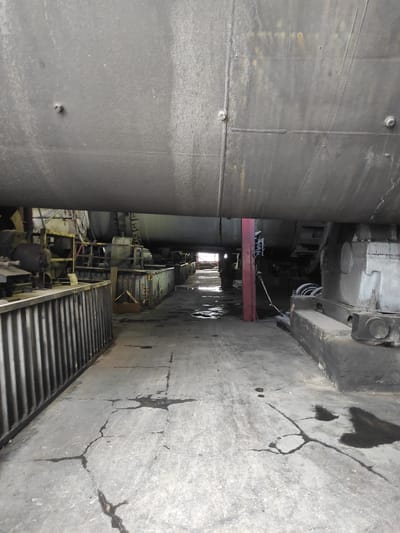 The image shows a dimly lit industrial setting, likely a maintenance or storage area, beneath a large, dark, cylindrical structure. The ground is cracked concrete, with several dark, wet patches indicating spills or leaks. To the left, a corrugated metal barrier runs along the edge of the scene. In the background, industrial machinery and equipment are visible, including metal structures, pipes, and what appear to be large rotating drums or tanks. A person in dark clothing is partially visible in the distance, standing near a doorway or opening, illuminated by light from beyond. The overall impression is of a functional, somewhat neglected industrial space. The specific location in Kyzyl, Kazakhstan, is not determinable from the visual information alone.