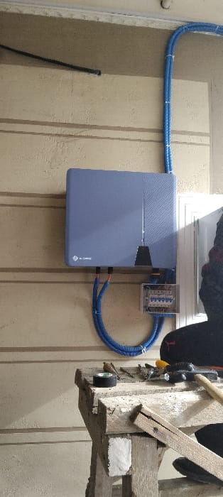 An electrical panel is mounted on a wall next to a blue rectangular box. The box has a two-toned design and a logo at the bottom right. Two blue conduits run along the wall, connecting to both the panel and the box. A window is positioned above the panel and box. To the right of the setup, a wooden structure with tools hanging from it is visible. The wall has a striped texture.