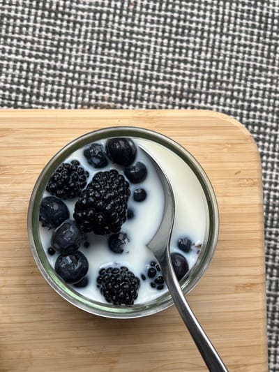 An overhead shot captures a glass container filled with a creamy white substance, likely milk, yogurt, or panna cotta, studded with fresh, dark berries. Several plump blueberries and two large blackberries are visible, some partially submerged in the liquid. A silver spoon rests within the container, its bowl partly submerged, suggesting it is being eaten or prepared for consumption.
The container is placed on a light-colored wooden cutting board or tray, exhibiting a distinct grain running horizontally. To the left, a portion of a black and white woven fabric, possibly a tablecloth or placemat with a small checkered or houndstooth pattern, forms the background.
The scene suggests a simple, healthy breakfast, dessert, or snack. There are no people visible, nor any discernible text. The lighting appears soft, indicating an indoor setting. This image is set in Aarhus, Denmark.
