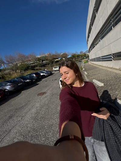 The image portrays an outdoor scene under a bright, clear blue sky in Porto Salvo, Portugal. A woman occupies the foreground, positioned slightly to the right, extending her right arm towards the camera, implying a selfie capture. She has brown hair, wears a maroon long-sleeved sweater over a white undershirt, light grey jeans, and carries a dark plaid jacket in her left arm. A brown braided bracelet is visible on her right wrist. Her expression is a smile accompanied by a wink of her left eye.

The background features an asphalt road curving uphill from the left foreground. Several vehicles are parked along the left side of the road, including a dark grey Tesla Model 3 (license plate 09 AA 69) and a white hatchback further in the distance. To the left of the road, an embankment is visible with sparse, bare trees and dry grasses. On the right side of the frame, a large, modern building with a curved concrete facade and horizontal architectural elements runs parallel to a paved sidewalk. Concrete blocks line the edge of the sidewalk. A lone individual is discernible walking uphill in the far distance on the road. Strong sunlight illuminates the scene, creating distinct shadows from subjects and objects.