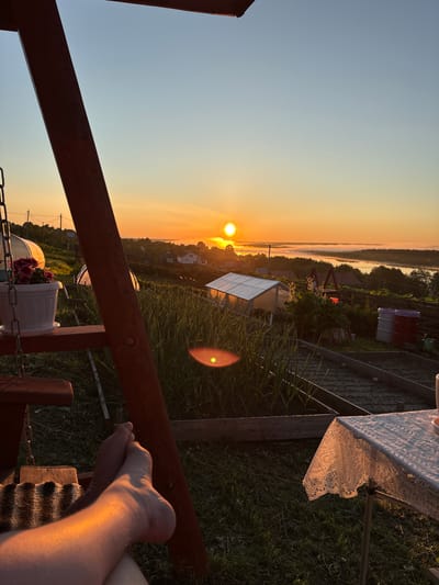 This image captures an outdoor rural scene at either sunrise or sunset in Зеленец, Russia. The sun, a bright orange orb, is low on the left horizon, illuminating the landscape with warm, golden light. The sky transitions from a deep orange at the horizon to a clear light blue overhead. In the distant background, a body of water, likely a river, is visible, partially obscured by a layer of low-lying fog or mist, with a band of dark trees lining its far bank.
The foreground is dominated by a dark wooden structural beam, possibly part of a swing set or pergola, which angles from the bottom left towards the top right. A person's bare feet and lower legs are visible in the bottom left, resting on a textured surface, strongly illuminated by the sun. On the left, a white pot containing pink flowers sits on a wooden ledge of the structure.
The midground features a cultivated garden area with visible rows of green plants, likely in raised beds, and several transparent or white-sheathed greenhouses/polytunnels. One large greenhouse is prominent in the mid-left. Two large barrels, one red and one white/purple, are situated near a wooden fence in the mid-right. Scattered houses and power lines are visible on the distant slopes. On the far right edge, a table covered with a white lace tablecloth is partially visible. A bright lens flare is present in the lower-right quadrant of the image.