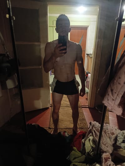 The image is a full-length, low-angle, indoor mirror selfie. A shirtless man is standing in what appears to be a hallway or doorway. He is holding a smartphone in his right hand, taking the picture. He is wearing dark shorts with a lighter waistband. His arms are down by his sides, and his legs are slightly apart.
The environment is dimly lit and appears to be a domestic interior. To the left of the man, a tall, dark mirror is visible, reflecting part of the scene. Behind him, a doorway leads into another room with orange-toned doors and a light fixture on the ceiling. To the right of the man, laundry is piled on the floor, including a pink garment with a star pattern. A rug with an intricate pattern is on the floor.
The scene does not contain any specific landmarks or details that would allow for definitive identification of the location as Fastiv, Ukraine. However, the overall impression is of a cluttered, lived-in space within a residential building.