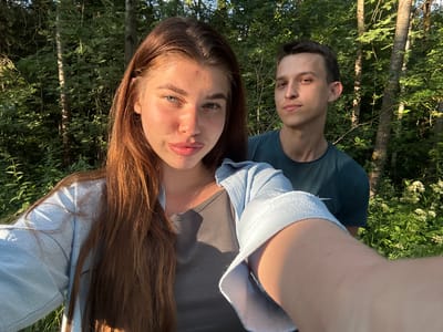In this outdoor, handheld selfie taken in a wooded area, a young woman with long, auburn hair and a light blue open-front shirt over a gray tank top is in the foreground, looking towards the camera. Her skin is flushed, suggesting sunlight exposure. To her right and slightly behind her, a young man in a dark teal t-shirt looks towards the camera with a neutral expression. The background consists of dense green foliage and trees, indicating a natural, possibly rural, environment. The lighting suggests it is late afternoon or early evening, with warm sunlight filtering through the trees. The location is identified as Шевлягино, Russia.