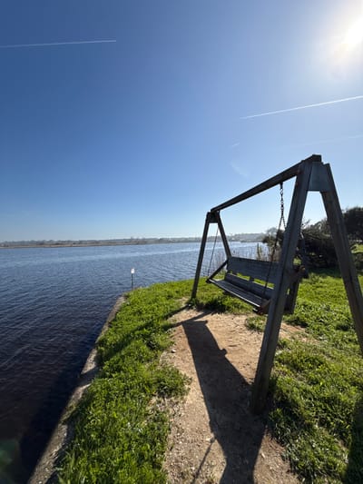 The image depicts a static outdoor scene in Praia de Mira, Portugal, under clear, bright sunlight. In the foreground, positioned to the right, a weathered wooden swing set with an empty wooden bench seat is present. The swing structure features two A-frame supports connected by a horizontal beam, from which the bench hangs via metal chains. A distinct, elongated shadow from the swing set extends across the light brown dirt and sparse green grass on the ground. To the left, a calm body of dark blue water with subtle ripples is visible, bordered by a concrete or stone embankment. A small white buoy or marker on a stick is partially submerged near the water's edge in the mid-ground. Beyond the water, a distant shoreline with faint green vegetation and structures extends towards the horizon. The sky, occupying the upper portion of the frame, is a brilliant, clear blue. The sun is visible in the upper right, casting a bright flare, and two distinct white contrails traverse the sky. No individuals, animals, or active interactions are present in the scene.