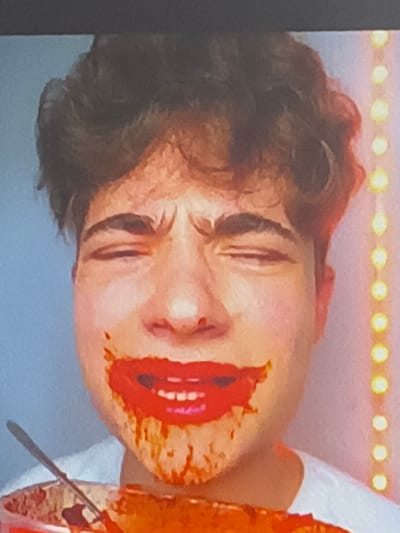 The image depicts a person with light skin and brown hair. Their eyes are closed and their face is tilted towards the right. Bright red substance is smeared around their mouth, resembling exaggerated lipstick. Part of a white garment is visible at the upper right of the frame. A small rod or stick is located nearby. The scene appears to be artificially lit, possibly a studio environment, indicated by the blurred lights at the top of the image. The image was captured indoors. There is no information on the location in the city of Kubrat, Bulgaria.