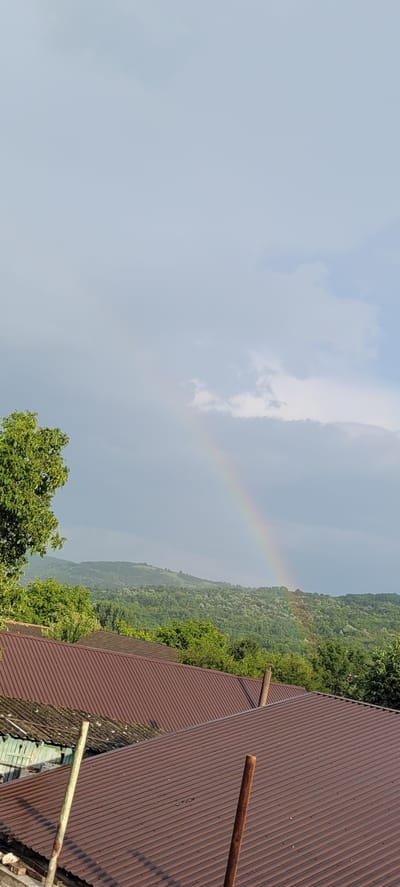 A post-rain landscape is depicted, featuring a faint rainbow arching across a cloudy sky over lush green hills. In the foreground, several reddish-brown corrugated metal roofs of houses are visible. A large green tree with dense foliage is on the left side of the frame. The scene appears to be in or around Zubrești, Moldova, suggesting a rural or semi-rural setting. There are no people or animals present in the image. The overall atmosphere is serene, with the natural beauty of the landscape enhanced by the presence of the rainbow.