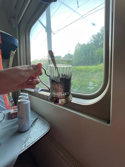 The image shows a hand holding a glass of water in a train compartment. The glass is in a metal holder with an ornate design. Outside the window, trees and green fields are visible, suggesting a rural or suburban setting. There are two cans and a small box on the table. The interior of the train compartment is visible, with beige and brown tones. The photo appears to be taken from inside the train, looking out the window. The image does not provide specific clues to locate it in the city of Keramik, Russia.