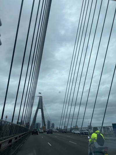 The image depicts a multi-lane road on the Anzac Bridge in Pyrmont, Sydney, Australia, viewed from inside a vehicle. The perspective looks along the bridge towards its main V-shaped concrete pylon, which is topped by a flagpole displaying an Australian flag. Numerous black suspension cables extend diagonally upwards from the bridge deck, framing the central pylon against an overcast, grey sky.

Vehicular traffic occupies the bridge deck. In the foreground, slightly right of center, a person on a dark motorcycle is visible from the rear. The rider is wearing a bright yellow-green high-visibility jacket, a dark helmet, and a black backpack. The motorcycle's rear license plate is partially discernible. Multiple cars and a truck are observed further along the bridge in various lanes, traveling in the same direction as the camera. Some vehicles are stationary or parked along the right side of the bridge deck. The cityscape, including the Sydney Tower, is visible in the far background to the left of the bridge. A circular smudge or glare is present on the vehicle's windshield above the central pylon. The scene is illuminated by ambient light under heavy cloud cover.