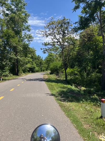 The image captures a paved road with a yellow dashed line down the center, winding through a lush green forest. The road is bordered by dense vegetation, including trees and shrubs, under a bright blue sky with scattered white clouds. In the foreground, a circular mirror reflects a person wearing a helmet and possibly riding a motorcycle. The scene appears to be on a sunny day, likely in a rural or natural area. The red and white marker post on the right side of the road suggests it might be a path or a less-trafficked route.