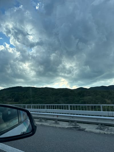 This is a photograph taken from the perspective of a moving vehicle, likely from the driver's or passenger's seat, on a bridge. The scene captures a cloudy sky with patches of blue visible, looming over a dense, dark green forested hillside. In the foreground, a silver guardrail runs along the side of the bridge, separating the roadway from the drop below. The asphalt road surface is visible in the lower portion of the image, with white lane markings on the far left. A car's side mirror is visible on the left side of the frame, reflecting a fragmented image of the sky and possibly the interior of the car. The location is identified as Dolno Varshilo, Bulgaria.