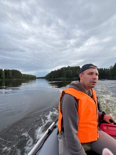 In a medium shot, a Caucasian man wearing an orange life vest and a dark baseball cap is seated in an inflatable boat, looking to the right of the frame. He has light skin and short, dark hair. The boat is moving, creating a wake in the dark water. The background shows a wide river flanked by dense green forests under a cloudy sky. The scene appears to be on a river in or near Anteplevo (Okhotinskoe S/P), Russia. The man's right hand is visible, holding a tiller or control lever. A red object, possibly a fuel can, is visible to his right.