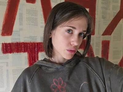 A medium shot depicts a young adult female positioned indoors, looking directly forward towards the camera. She has a light complexion, light blue-green eyes, and medium brown hair styled in a bob, parted on her left side. Her expression is neutral with slightly parted lips. She is wearing a dark grey, faded hooded sweatshirt with a distressed red floral or abstract design centered on the chest. The background is a wall covered with numerous sheets of light-colored paper, resembling aged book pages, displaying printed text in columns. Overlaid on these papers are broad, textured applications of red paint, forming abstract shapes. One prominent red shape, partially visible behind her head and left shoulder, resembles a large capital 'A'. Another red horizontal bar with textured details is visible behind her right shoulder. The setting is an indoor environment. Location: Doral, United States of America.